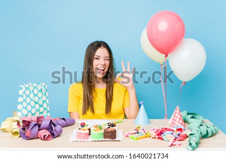 Young caucasian woman organizing a birthday winks an eye and holds an okay gesture with hand.