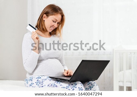A pregnant woman is sitting on a bed using a laptop and holding a plastic Bank card. The concept of online shopping and modern technologies. Close up