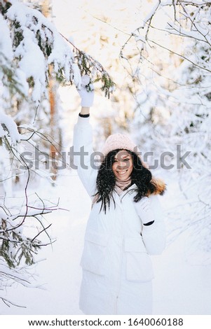 Outdoor close up portrait of young beautiful happy smiling girl wearing white knitted beanie hat, scarf and gloves. Model posing in park with Christmas lights. Winter holidays concept.