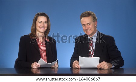 Male and female newscasters sitting at desk smiling at viewer. Royalty-Free Stock Photo #164006867