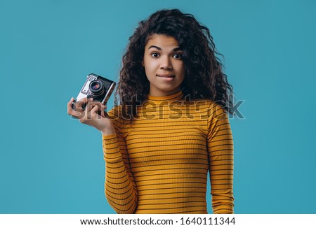 Beautiful african american girl dressed in striped orange golf holds retro camera in hand isolated on a blue background.