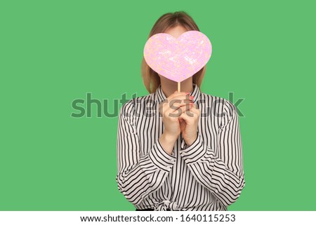 Anonymous dating. Unknown woman in glamour striped blouse covering face with pink heart, hiding behind symbol of love, romance on Valentine's day. indoor studio shot isolated on green background