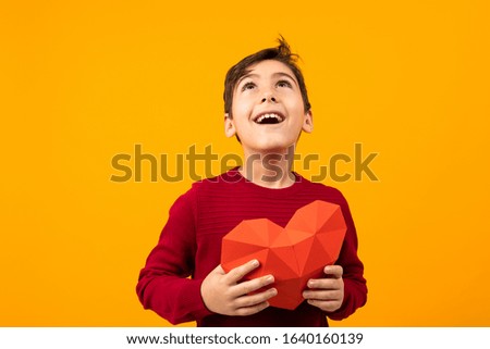 funny cheerful handsome caucasian boy with paper heart for valentines day on orange background