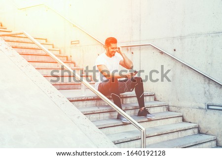 Side view of cheerful active male in sportswear adjusting earphones and choosing playlist on mobile phone while sitting on stairs and preparing for training in city