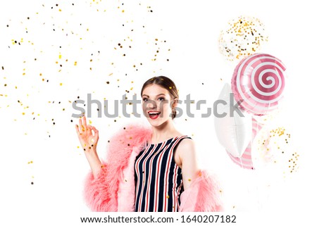 Funny woman celebrating with air balloons isolated white. Shows hand okay. in a pink fur coat, golden tinsel.Vivid emotions, surprise and joy, laughter and scream.Birthday holiday party.