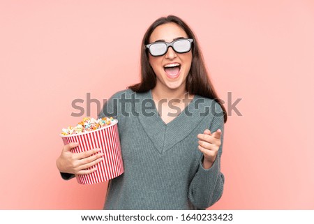 Young caucasian woman isolated on pink background with 3d glasses and holding a big bucket of popcorns while pointing front