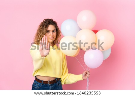 Young blonde woman with curly hair catching many balloons isolated on pink background making stop gesture with her hand