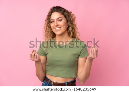 Young blonde woman with curly hair isolated on pink background making money gesture