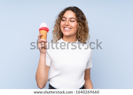 Young blonde woman with curly hair holding a cornet ice cream isolated on blue background with happy expression