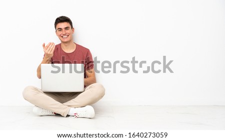 Teenager man sitting on the flor with his laptop inviting to come with hand. Happy that you came