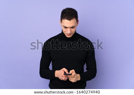 Young caucasian handsome man with turtleneck sweater isolated on purple background sending a message with the mobile