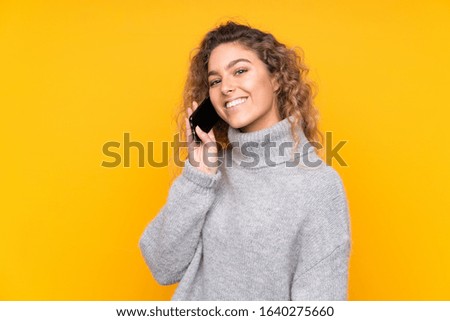 Young blonde woman with curly hair wearing a turtleneck sweater isolated on yellow background keeping a conversation with the mobile phone