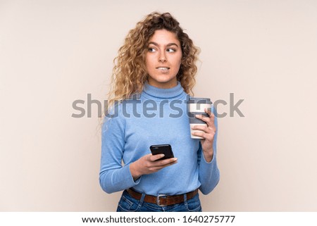 Young blonde woman with curly hair wearing a turtleneck sweater isolated on beige background holding coffee to take away and a mobile while thinking something