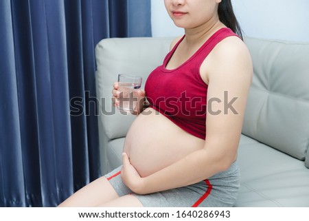 Beautiful asian pregnant woman wearing a red shirt while drinking water, Young woman  in livingroom.
