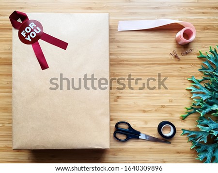A minimal brown paper gift box with wrapping accessories lay flat on wooden table on the center of the frame with palm leave. Close up, top view and isolated on wooden background, with copy space.