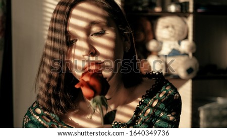 Portrait of beautiful girl sitting by window at sunset with beautiful flower of red rose. Valentine Day, march 8, February 14, birthday, anniversary, first date concept. Close up view