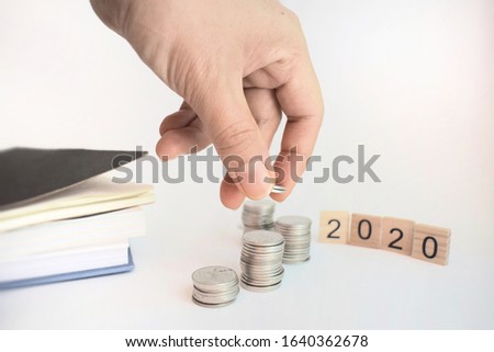 Financial matters. One hand is picking a coin from a pile of silver coins, on the left there are books or notebooks and on the right there are wooden blocks painted with numbers arranged as 2020.