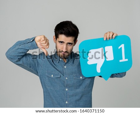 Attractive unhappy young man holding social media symbol of dislike feeling sad and rejected in internet obsession, social network notification icons, cyber bullying and technology communication.