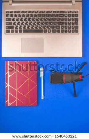 A microphone for recording sound and podcasts next to a laptop and notebook on a blue background. Desktop of blogger. Top view, flat lay.