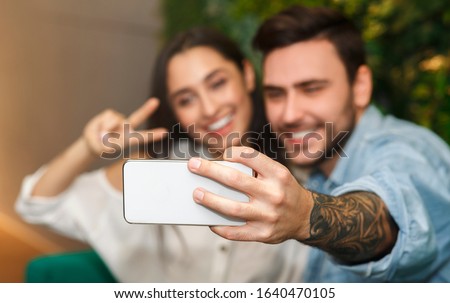 Happy Couple Making Selfie On Smartphone Gesturing V-Sign Having Fun During Romantic Date Sitting Together In Cafe. Selective Focus