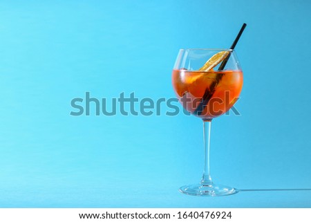 A glass of fruit liqueur in wine glass with a piece of lemon and black plastic straw isolated on pastel blue background. Summer alcohol drink. Copy space.