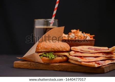 American snack, big burger, a glass of cola, slices of pizza and a bowl of popcorn. Tasty and appetizing food on black background. Fast food, junk food concept. Isolated on black with copy space.
