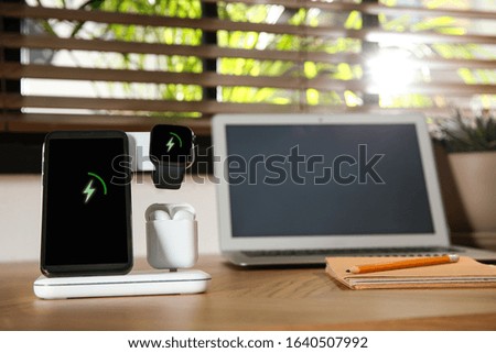 Mobile phone, earphones and smartwatch charging with wireless pad on wooden desk, space for text. Modern workplace device