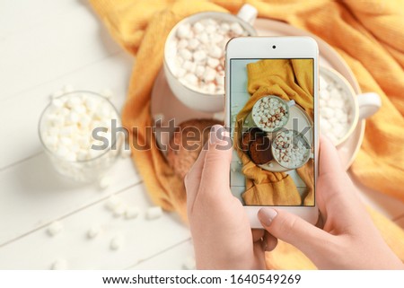 Female food photographer with mobile phone taking picture of hot chocolate with marshmallows