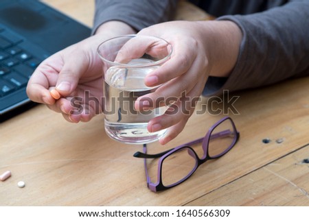 A hand with water tumbler and pills with specs and laptop.