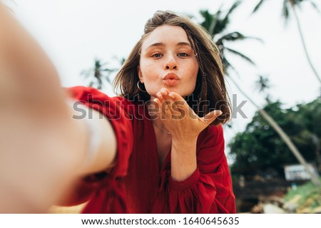 Young attractive woman in red dress send kisses to camera and hold it with one hand. Stand alone on beach with green palm trees beinhd. Love is in air. Female model posing. Resort on tropic island