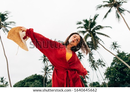 Playful positive young woman dancing alone and having fun. Low view of female model singing and looking on camera. White sky and green palm trees. Woman wear red beach dress