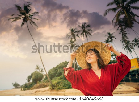 Picture of gorgeous young woman having rest in apradise island. Look up in sky and smile. Female model has rest on island or paradise beach. Wear red dress and beautiful hat. Sky getting darker