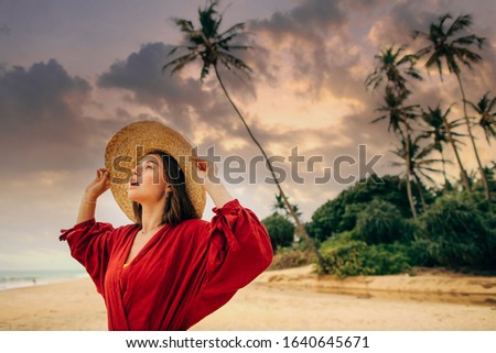 Young amazed woman look up on sky. Stand on beach sand on paradise island alone. Wear hat and red pareo or dress. Green palm trees and blue ocean. Emotional beautiful lady