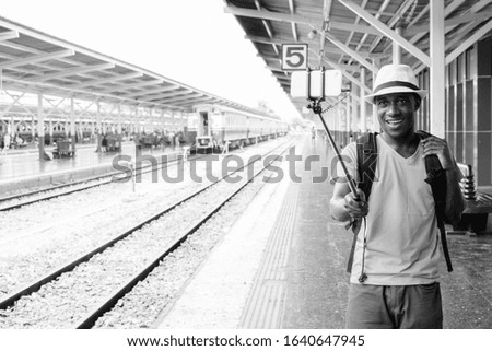 Young happy African tourist man taking selfie with phone on selfie stick at the railway station