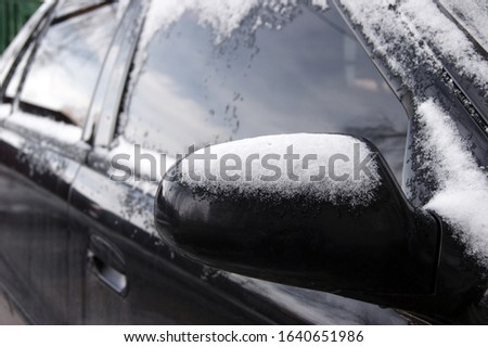 Side of a snowy black car with mirror on a cold winter morning, horizontally framed picture.