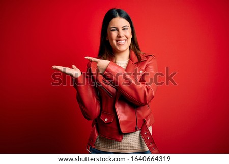 Young beautiful woman wearing red leather jacket over isolated background amazed and smiling to the camera while presenting with hand and pointing with finger.