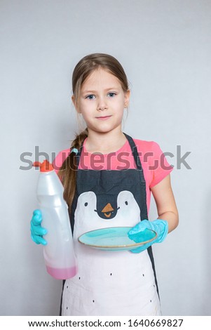A small cheerful European girl holds a dish detergent and a clean plate in her hands. The child helps in the kitchen to wash dishes.