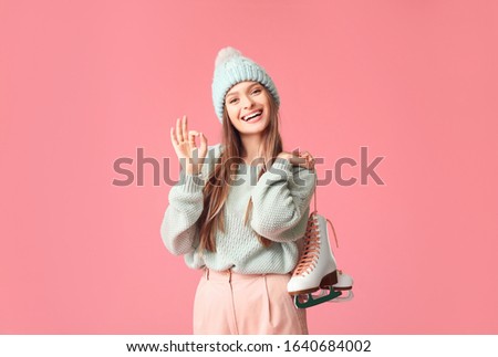 Beautiful young woman with ice skates showing OK on color background