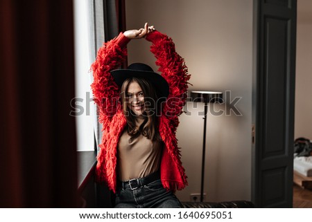 Merry lady with charming smile looks into camera against background of room with lamp. Girl in hat and woolly jacket raises hands