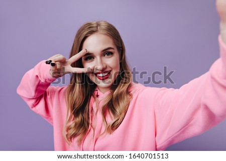Beautiful woman in pink hoodie showing peace sign and taking selfie on purple background. Happy girl in bright sweatshirt smiling widely on isolated