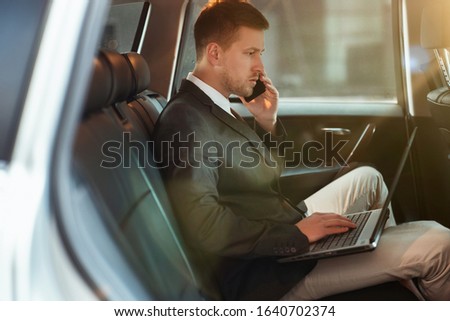 young businessman works in his laptop having business phone conversation while sitting in his car on his way to office, multitasking concept