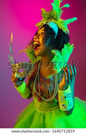 Close up. Beautiful young woman in carnival, stylish masquerade costume with feathers on gradient background in neon light. Concept of holidays celebration, festive time, dance, party, having fun.