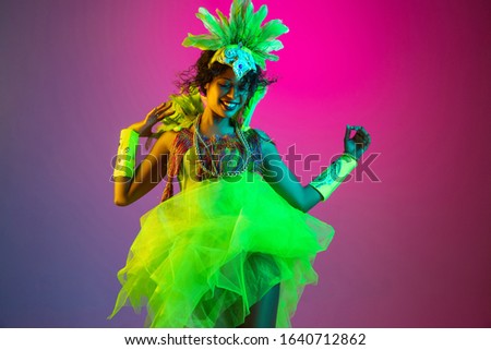 Colors. Beautiful young woman in carnival, stylish masquerade costume with feathers dancing on gradient background in neon light. Concept of holidays celebration, festive time, dance, party, having