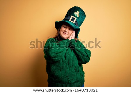 Young beautiful plus size woman wearing green hat with clover celebrating saint patricks day sleeping tired dreaming and posing with hands together while smiling with closed eyes.