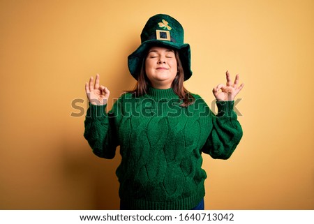 Young beautiful plus size woman wearing green hat with clover celebrating saint patricks day relax and smiling with eyes closed doing meditation gesture with fingers. Yoga concept.