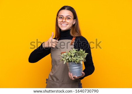 Young gardener girl holding a plant over isolated yellow background giving a thumbs up gesture