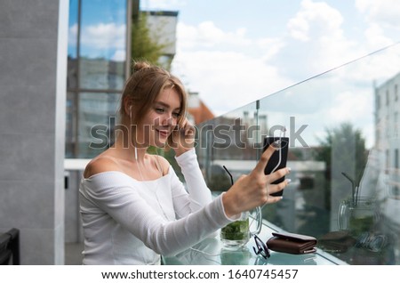 Photo of beautiful lady sitting in cafe and making selfie with a smart phone. Portrait of young smiling student girl make video call from smartphone to her best friend, having break, telling good news