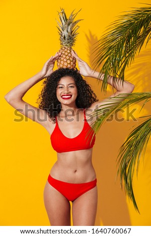 Portrait of a beautiful happy young fit african woman wearing bikini standing isolated over yellow exotic background, posing with pineapple