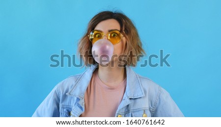Portrait happy smile woman in star sunglasses inflates bubble from chewing gum balloon and bursts it in on blue background. Emotions of people. Lifestyle. Party. Monotone. Blue pantone 