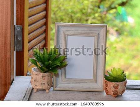 The cactus is in a clay vase in front of a picture frame on the table by the window in the room.